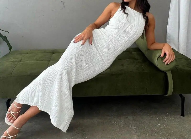 A woman sitting on a green bench wearing a white sleeveless maxi dress with a high neckline and ruffled hem.